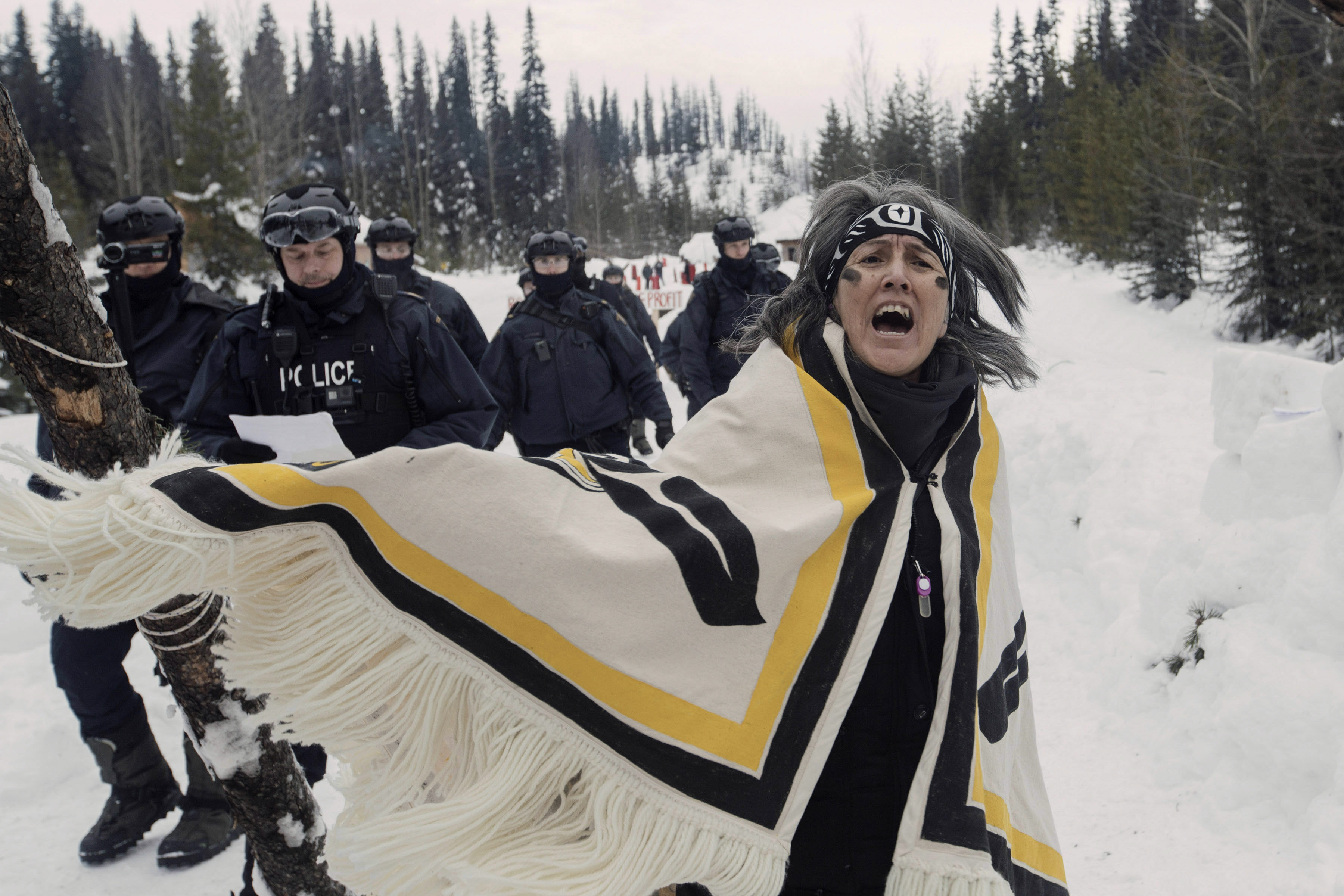 Against a snowy, tree-lined landscape, a woman with charcoal on her face sings and dances while police approach from behind.